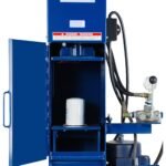Close-up of a blue industrial oil filter crusher with the door open, showing a white spin-on filter positioned inside the crushing chamber. Hydraulic hoses, gauge, and control lever are visible on the right side of the machine