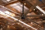 Cool Boss CB-24 HVLS 24-foot industrial ceiling fan mounted in a high-bay workshop with string lighting and exposed rafters, providing wide-area air circulation.