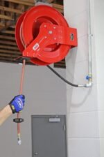 Technician pulling hose from a wall-mounted Lincoln 83754 Value Series reel, showing ergonomic use in an auto repair or industrial workspace. Get it at Wrenchers.com